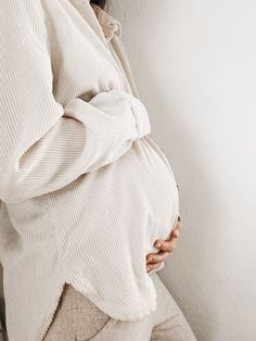 a pregnant woman wearing a white sweater and beige pants, leaning against a wall with her hands on her stomach