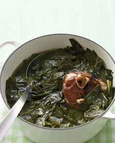 a pot filled with greens and meat on top of a green table cloth next to a white spoon