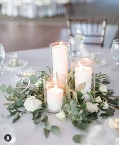 candles are lit on a table with greenery and white roses in front of it