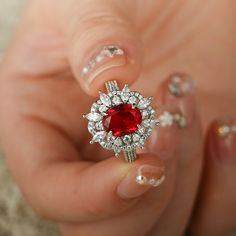 a woman's hand holding a ring with a red stone surrounded by white diamonds