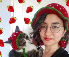 a woman wearing glasses and a knitted strawberry hat holds a cat in her arms