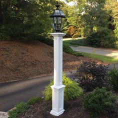 a white lamp post in the middle of a park with trees and bushes around it