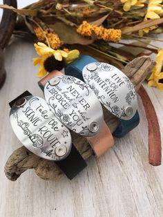 three leather bracelets with writing on them sitting next to yellow flowers and dried twigs