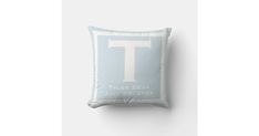 a light blue pillow with the letter t printed on it, sitting against a white wall