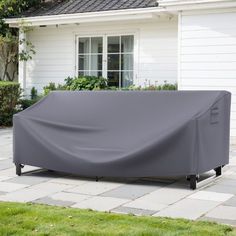 an outdoor couch covered in a gray cover sitting on top of a brick patio next to a white house