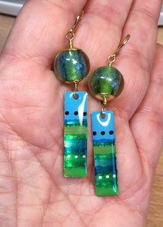 Springy Greens These earrings are created from artisan made glass beads. The round beads have a core of spring green, with a net-like coat of blue over them. They are encased with a thick coat of clear glass which magnifies the color and texture. Below them dangle artisan brass charms, 1 1/8 inch long and 3/8 inch wide. They have been painted with irregular stripes in a variety of blues and greens, both flat and sparkly, with dots of black and gold, then sealed with a thick coat of resin. I have Artsy Green Drop Earrings, Artistic Green Nickel-free Jewelry, Green Artsy Drop Earrings, Handmade Artsy Blue Beaded Earrings, Green Metal Beaded Drop Earrings, Nickel-free Green Metal Beaded Earrings, Artsy Blue Beaded Earrings, Artsy Green Earrings For Gift, Green Artisan Enamel Earrings