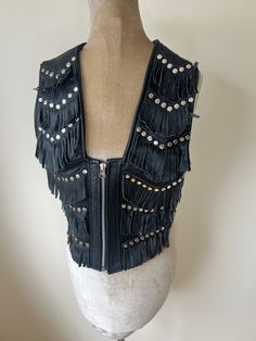 A vintage 1980's Nice London, black leather waistcoat with fringing and silver studs. It has straps and buckle fastenings at the side. Made from a nice soft leather. UK Size 8 - 10.  Please check measurements: Chest - 88cm Waist - 82cm Length - 46cm Gothic Vest For Fall Festival, Gothic Festival Vest For Fall, Edgy Black Vest For Festival, Edgy Black Festival Vest, Biker Style Sleeveless Vest For Festivals, Biker Sleeveless Vest For Festivals, Fitted Fringe Vest For Festival, Fitted Fringe Vest For Festivals, Leather Waistcoat