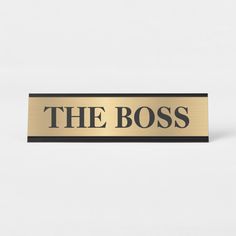 a sign that says the boss in black and gold on a white background with a shadow