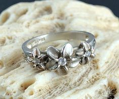 This is a lovely estate sterling silver stackable ring featuring three plumeria flowers with cubic zirconia CZ stones and etched shank. It is hallmarked 925 and tests positive for sterling silver with the 18K acid solution - a video of testing is available upon request. The gemstones were confirmed with the Presidium Gem Tester II - the video of testing is available upon request. Signed PCU. Weight: 2.53 g Size: 6-6 ¼ Width: 0.32" (0.82 cm) Very good pre-owned condition; please see photos and video. A precious metal polishing cloth is included. All jewelry is sold in used, pre-owned condition. Vintage and antique items are expected to have some wear, imperfections, abrasions, scuffs, signs of use and age. Please look carefully at the photos; they are part of the item description. Some imag Sterling Silver Stackable Rings, Botanical Theme, Plumeria Flowers, Hawaiian Flower, Black Patch, Hawaiian Flowers, Stackable Ring, Pretty Gift, Cz Stone