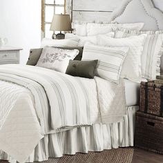 a bed with white and grey comforters in a bedroom next to a lamp on a dresser