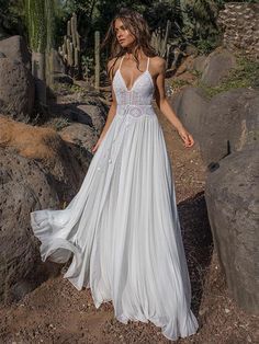 a woman in a white wedding dress standing on rocks near trees and cactuses with her back to the camera