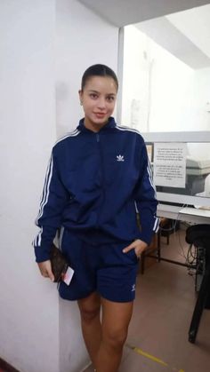 a woman standing next to a white wall wearing blue shorts and adidas jacket with her hands in her pockets