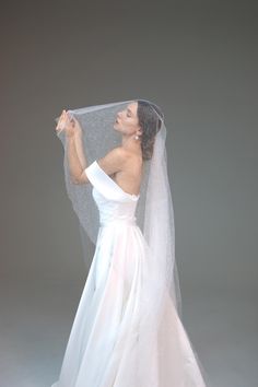 a woman in a white wedding dress holding a veil over her head and looking up