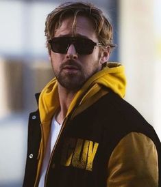 a man wearing sunglasses and a yellow hoodie looks at the camera while standing in front of a building