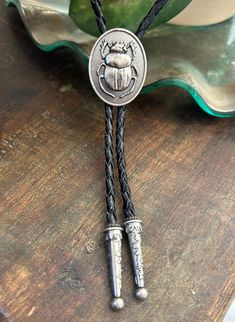 This awesome bolo tie has a meal beetle pendant. Quality guaranteed, picture does not do this buckle justice! The pendant piece measures 1 1/8'' x 1 1/4'' It can adjust lengths, 39'' in total length. The cord is genuine black woven leather.   We have matching belt buckles to pair! Men’s Leather Jewelry, Classic Silver Bolo Tie As Gift, Vintage Black Bolo Ties For Gift, Vintage Black Bolo Ties As Gift, Handmade Silver Bolo Tie As A Gift, Handmade Silver Bolo Tie For Gift, Silver Bolo Ties As A Gift, Brown Bolo Ties As A Gift, Brown Bolo Tie For Gift