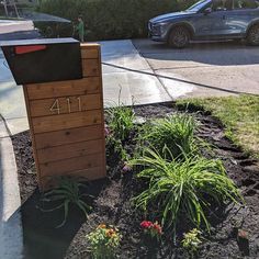 a mailbox sitting in the middle of a flower bed next to a sidewalk with cars parked on it