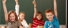 several children sitting at desks with their hands in the air