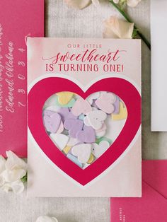 a greeting card with hearts and flowers on the table next to it is a pink envelope that says our little sweetheart is turning one