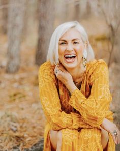 an older woman with white hair sitting on the ground in front of trees and smiling