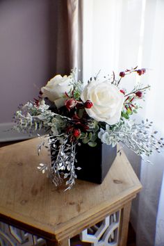 a black vase with white roses and greenery sits on a wooden table in front of a window