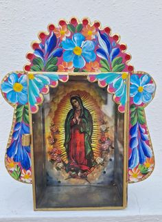 an image of the virgin mary with flowers in it's frame on a shelf