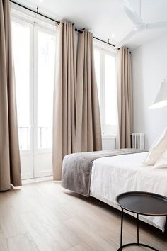 a white bed sitting next to a window with curtains on top of it and a table in front of it