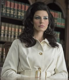 a woman sitting at a table in front of bookshelves wearing a white coat