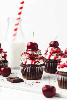 chocolate cupcakes with cherries and whipped cream are on a white tray next to a glass of milk