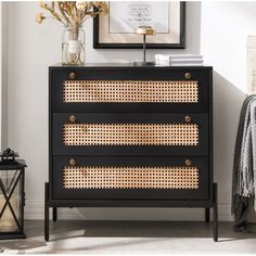 a black dresser with rattan drawers and gold handles in a white room next to a framed print on the wall