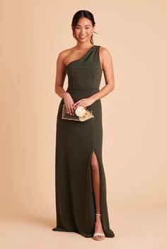 a woman in a one shoulder dress holding a clutch and smiling at the camera while standing against a beige background