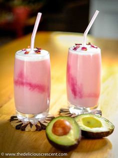 two glasses filled with fruit and drink on top of a wooden table next to an avocado