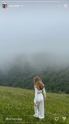 a woman standing on top of a lush green field next to a forest covered hillside