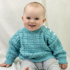 a baby sitting on top of a white sheet wearing a blue sweater and grey pants