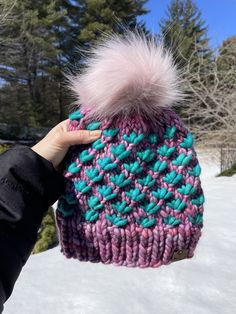 a person holding up a pink and blue knitted hat with a fuzzy pom