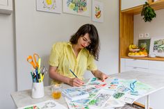 a woman is sitting at a table with some art work on it and holding a pencil