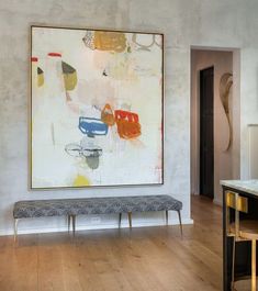 a painting hangs on the wall above a bench in a room with hardwood floors and white walls