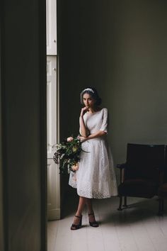 a woman in a white dress is standing by a door and holding a flower bouquet