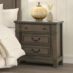 a night stand with two drawers and a lamp next to it on top of a bed