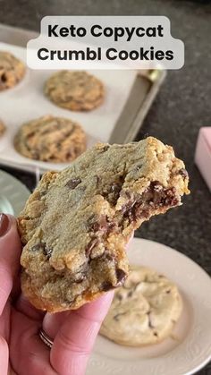 a person holding up a chocolate chip cookie in front of some muffins on a plate