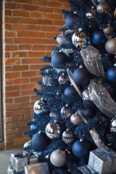 a blue christmas tree with silver and black ornaments on it, next to a brick wall