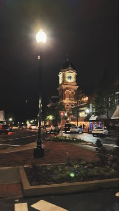 a clock tower is lit up at night in the middle of a town square with cars parked on the street