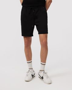 A man in Psycho Bunny eco-friendly shorts and white sneakers with black accents, standing on a plain background. The focus is on the lower half of his body from the waist down. Casual Black Stretch Athletic Shorts, Black Relaxed Fit Shorts With Short Inseam, Black Knee-length Loungewear Athletic Shorts, Black Knee-length Athletic Shorts For Loungewear, Black Knee-length Athletic Shorts For Lounging, Casual Black Fitted Athletic Shorts, Black Fitted Casual Athletic Shorts, Black Relaxed Fit Cotton Athletic Shorts, Black Relaxed Fit Knee-length Shorts