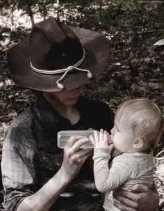 a woman with a cowboy hat holding a baby in her lap while sitting on the ground