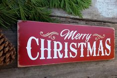 a merry christmas sign hanging on a wooden wall next to pine cones and evergreen branches