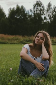 a woman is sitting in the grass with her hand on her head and looking at the camera