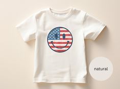 Introducing our Toddler 4th Of July American Flag Smiley T-Shirt, made for Comfort and Style!  :-) DESIGN: This toddler t-shirt features a smiley face colored in with the red, white and blue American flag, sure to show off their love for America! :-) VIBRANT COLORS: Made with high-quality, vibrant ink, the colors on this t-shirt are of exceptional quality and will not disappoint. *{Please note that due to variations in screen resolutions and printing processes, colors may slightly vary from what is displayed on your device. We strive to ensure accuracy in our product images, but slight differences in hue may occur. Rest assured, the charm and vibrancy of this item remains consistent, adding a delightful touch to your wardrobe.} :-) COMFORTABLE FIT: The Bella+Canvas 3001T t-shirt is made of Cute Cotton T-shirt For 4th Of July, Playful T-shirt For 4th Of July With Crew Neck, Playful Crew Neck T-shirt For 4th Of July, Cute American Flag Print T-shirt For Summer, Cute American Flag Print Short Sleeve T-shirt, Cute Independence Day T-shirt With Graphic Print, Cute Independence Day Graphic Print T-shirt, Cute Independence Day Graphic T-shirt, Playful White T-shirt For 4th Of July