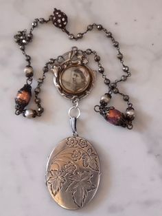 A delicate memento Mori assemblage featuring an Art Nouveau pendant from France suspending a British sterling locket with a floral chased design. The French pendant has what looks like a photo of a Normandy woman. I think the frame is silver plated, it is an antique dating around 1900. The locket is vintage. It has the interior brass bezels and plastic covers for photos. I have not taken these out to clean and there is white haze on the interior plastic. The chain is handmade pyrite rosary chain interspersed with Czech glass pearls and two antique Czech opal glass beads. Connects with a magnetic clasp. Handmade Antique Silver Vintage Locket Necklace, Handmade Vintage Locket Necklace In Antique Silver, Artisan Silver Jewelry With Vintage Charm, Ornate Handmade Silver Locket Necklace, Ornate Silver Handmade Locket Necklace, Handmade Antique Silver Pendant Locket Necklace, Handmade Victorian Jewelry Keepsake, Handmade Antique Silver Locket Necklace, Handmade Vintage Silver Locket Necklace