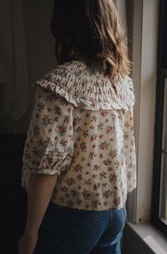 a woman is looking out the window with her back turned to the camera while wearing jeans and a floral blouse