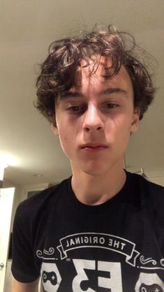 a young man with curly hair wearing a black t - shirt and looking at the camera