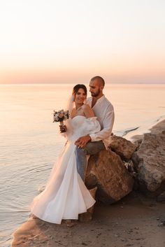 a bride and groom sitting on rocks at the edge of the water with sunset in the background